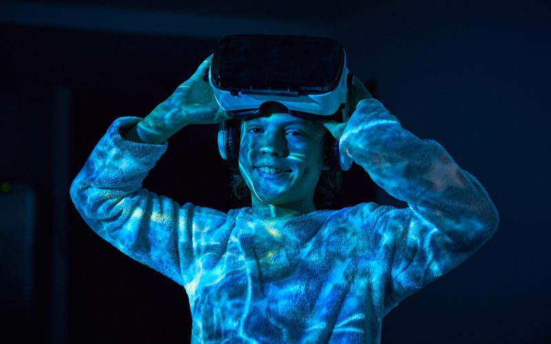 Cheerful little boy smiling and looking at camera while taking off VR goggles with headphones after playing video game in dark room